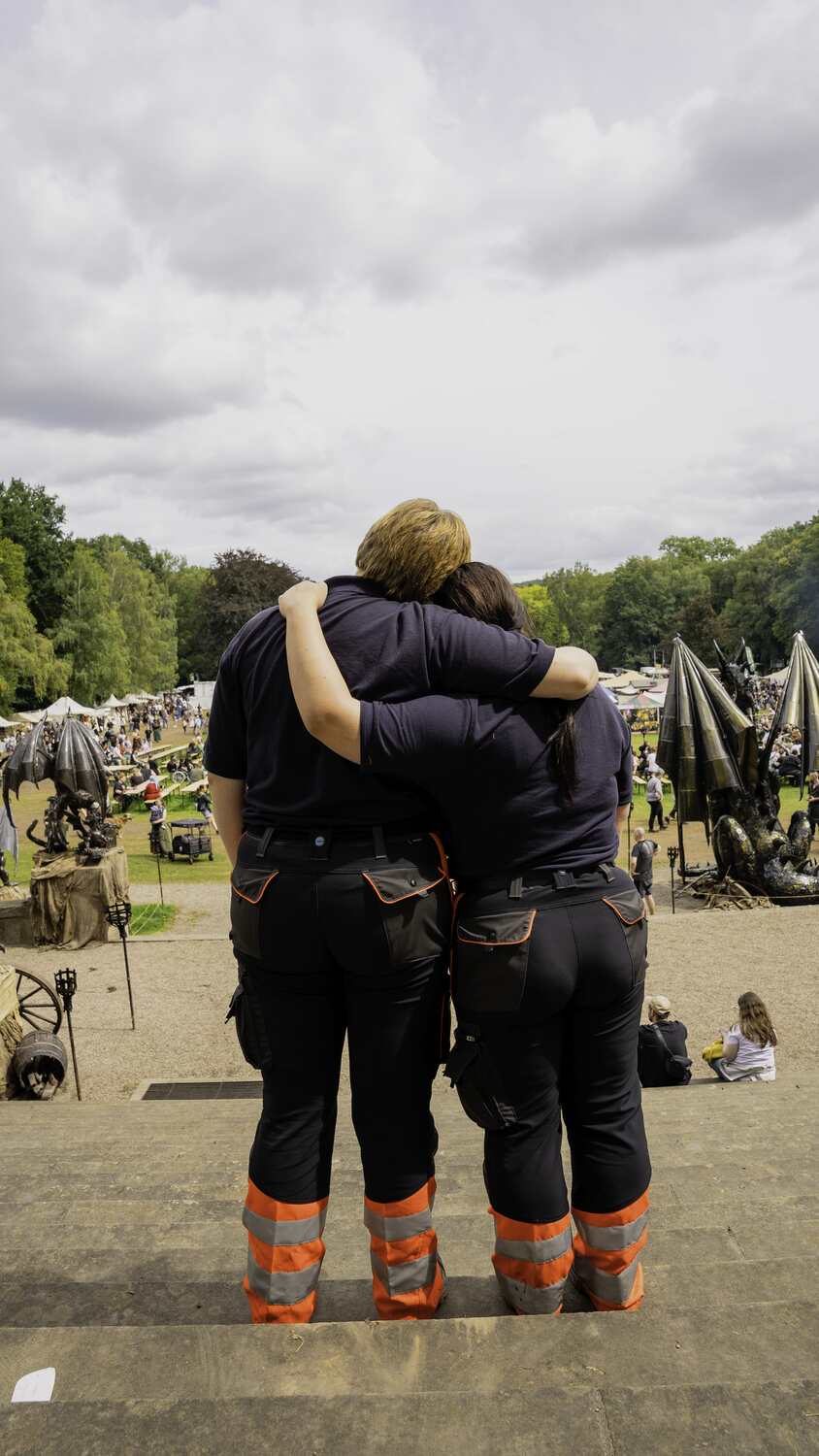 Zwei Personen in schwarzen Uniformen mit orangenen Reflexstreifen umarmen sich und blicken auf ein Outdoor-Event.