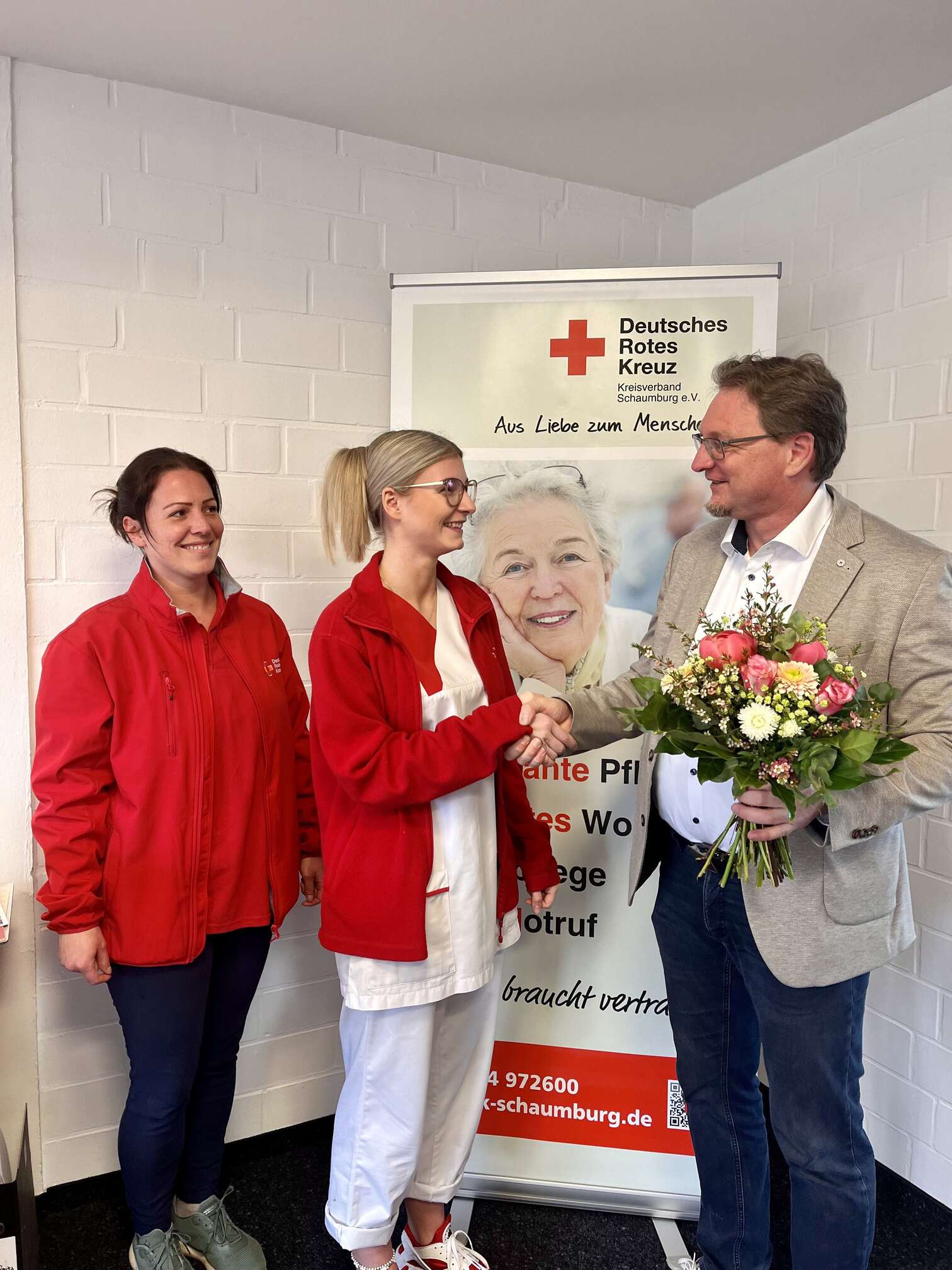 Mann im beigen Mantel überreicht Blumen und schüttelt einer DRK-Mitarbeiterin die Hand, eine weitere Kollegin steht daneben.
