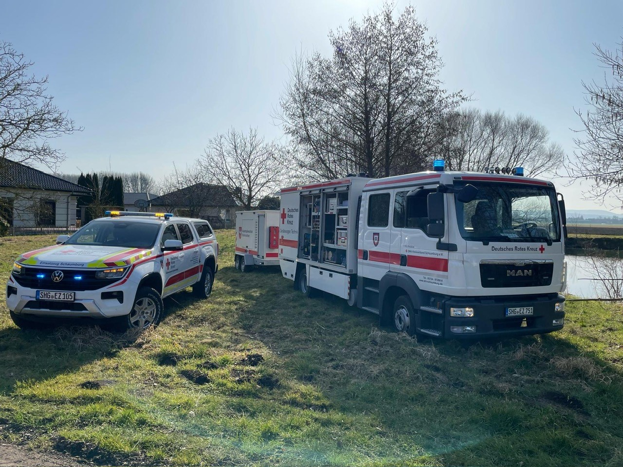 Weißes Volkswagen-Einsatzfahrzeug und MAN-Lkw mit Rotkreuz-Markierungen stehen auf einer Wiese nahe blattlosen Bäumen.