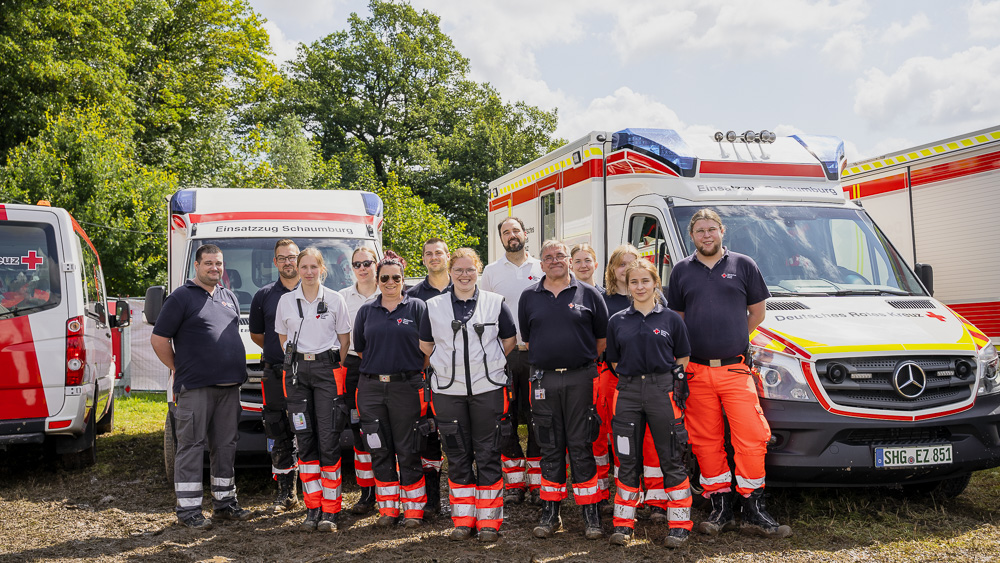 Gruppe von 13 DRK-Einsatzkräften steht vor drei Rettungswagen im Freien, im Hintergrund Bäume.