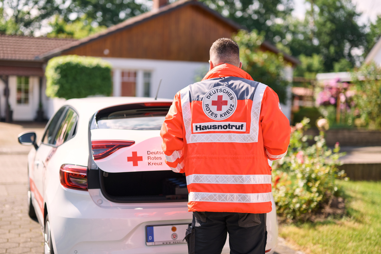 Hausnotruf-Einsatzkraft in leuchtend oranger DRK-Einsatzjacke steht hinter einem weißen Auto mit offenem Kofferraum in einem Wohngebiet. 