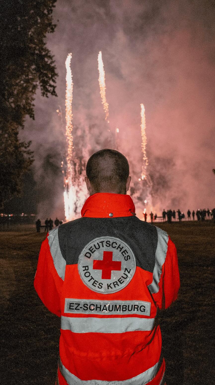 Person in einer DRK-Einsatzjacke steht nachts im Freien und schaut auf ein Feuerwerk in der Ferne.