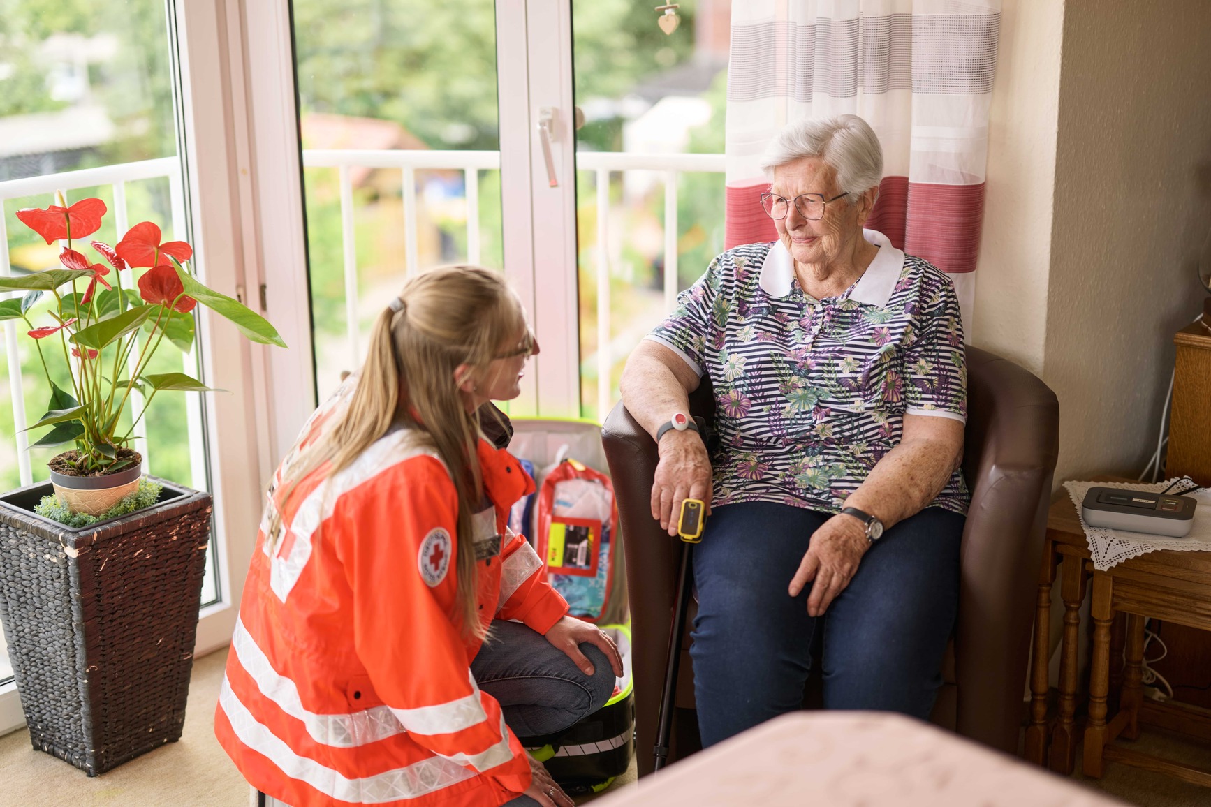 Ein DRK-Hausnotruf-Mitarbeiter in orangefarbener reflektierender Jacke kniet neben einer älteren Frau im Stuhl und hält ein Messgerät an ihrem Finger, in einem gemütlichen Raum mit Fenster und Topfpflanze.