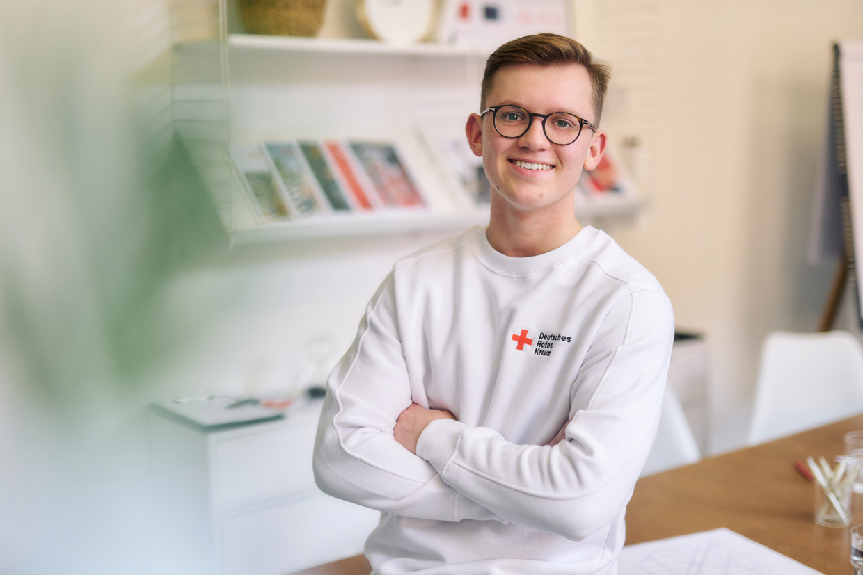 Lächelnder junger Mann mit Brille in einem weißen Deutsches-Rotes-Kreuz-Sweatshirt sitzt mit verschränkten Armen im Innenraum.