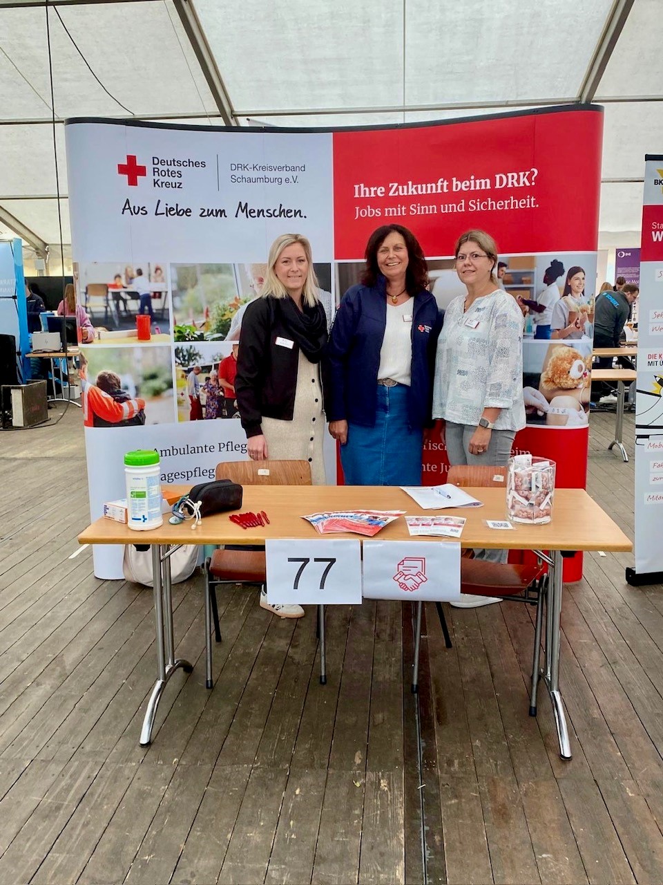 Drei Frauen stehen hinter einem DRK-Infostand mit Broschüren und einer Tischnummer 77.