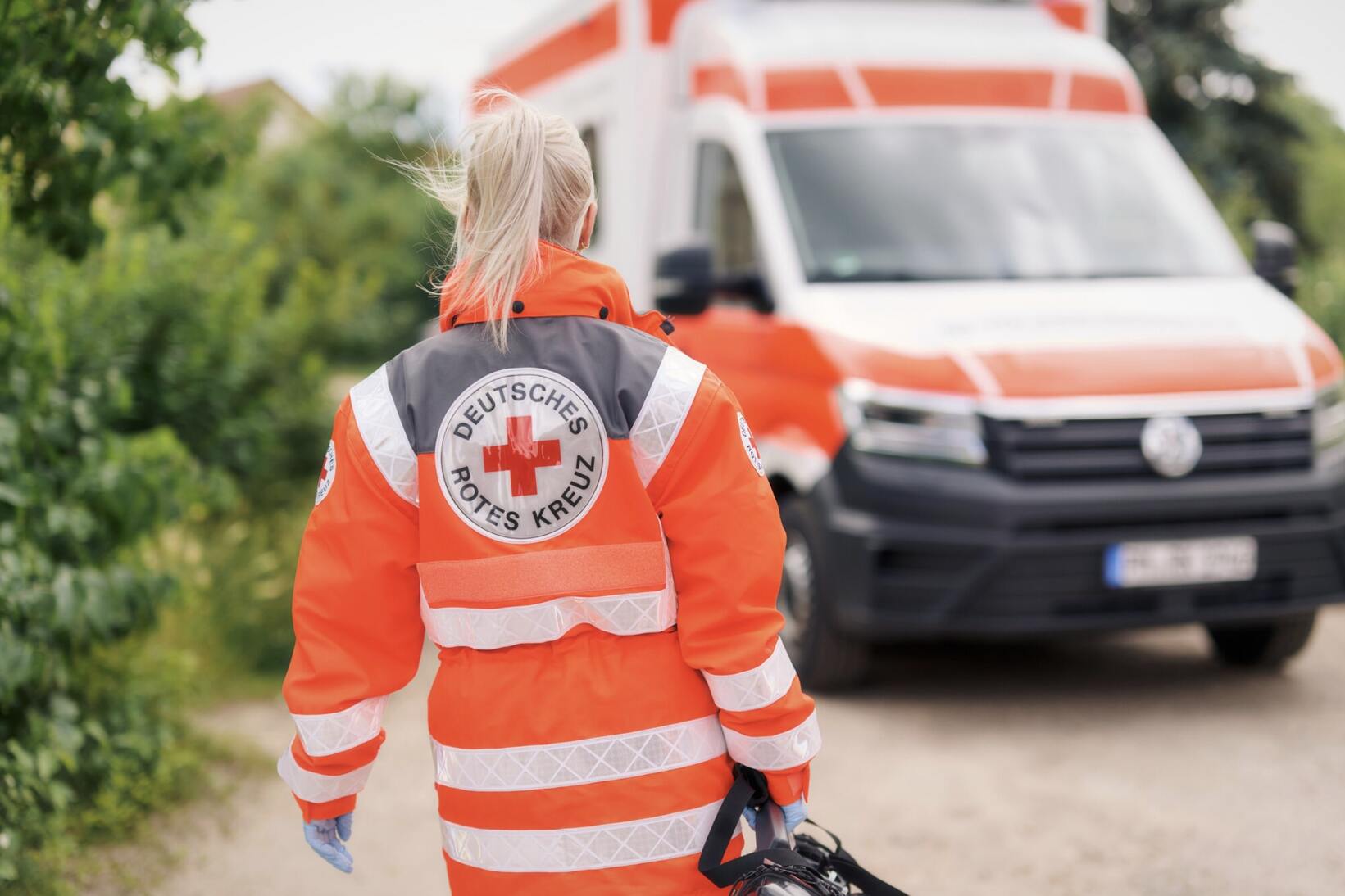 Rettungskraft in orangefarbener DRK-Uniform geht mit Helm in der Hand auf einen Krankenwagen zu.