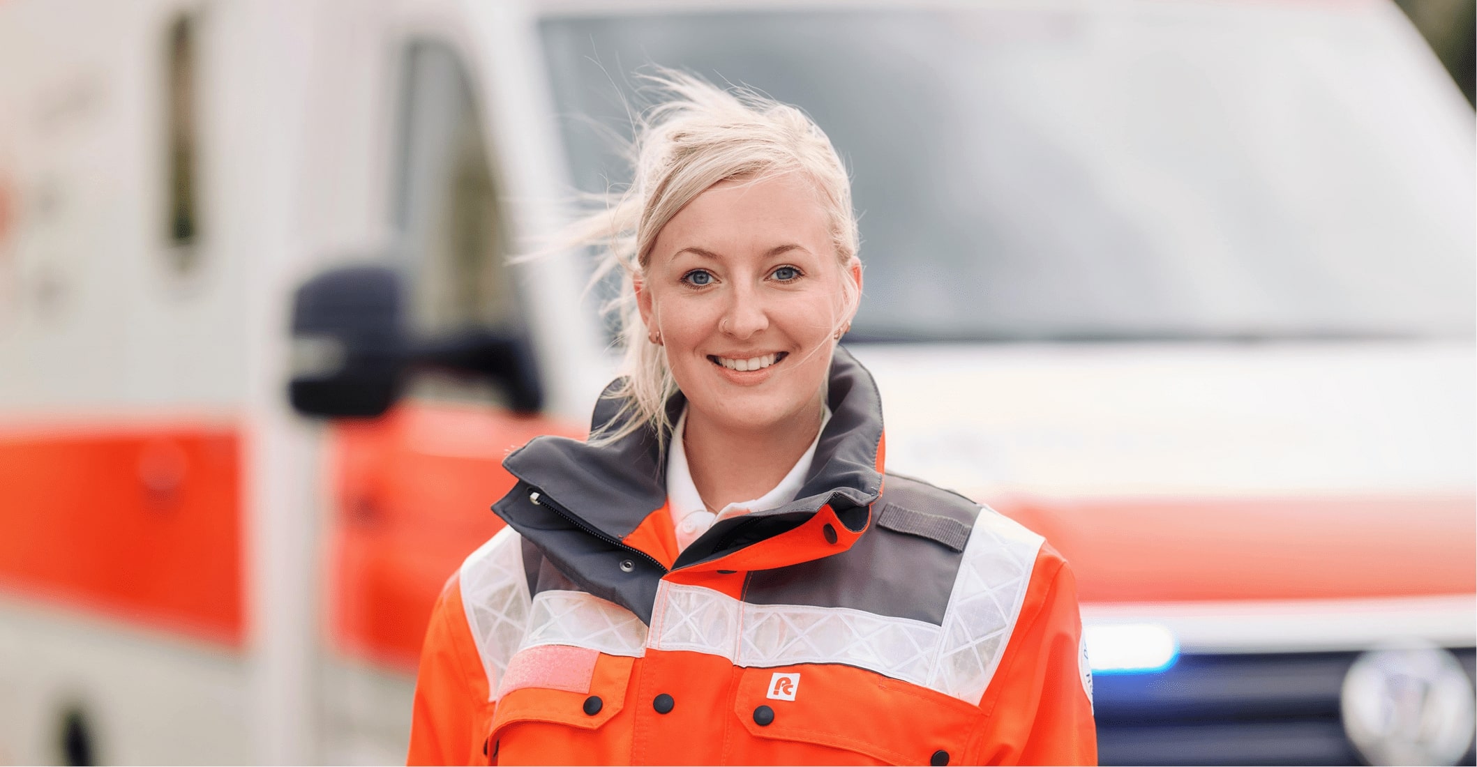 Lächelnde Rettungssanitäterin in orangefarbener Warnschutzkleidung steht vor einem Rettungswagen.