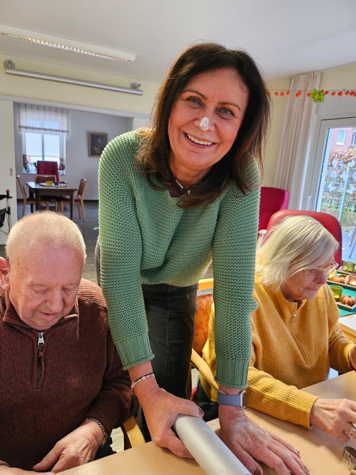 Lächelnde Frau mit Mehl auf der Nase lehnt an einem Tisch zwischen zwei älteren Menschen bei einer Aktivität.