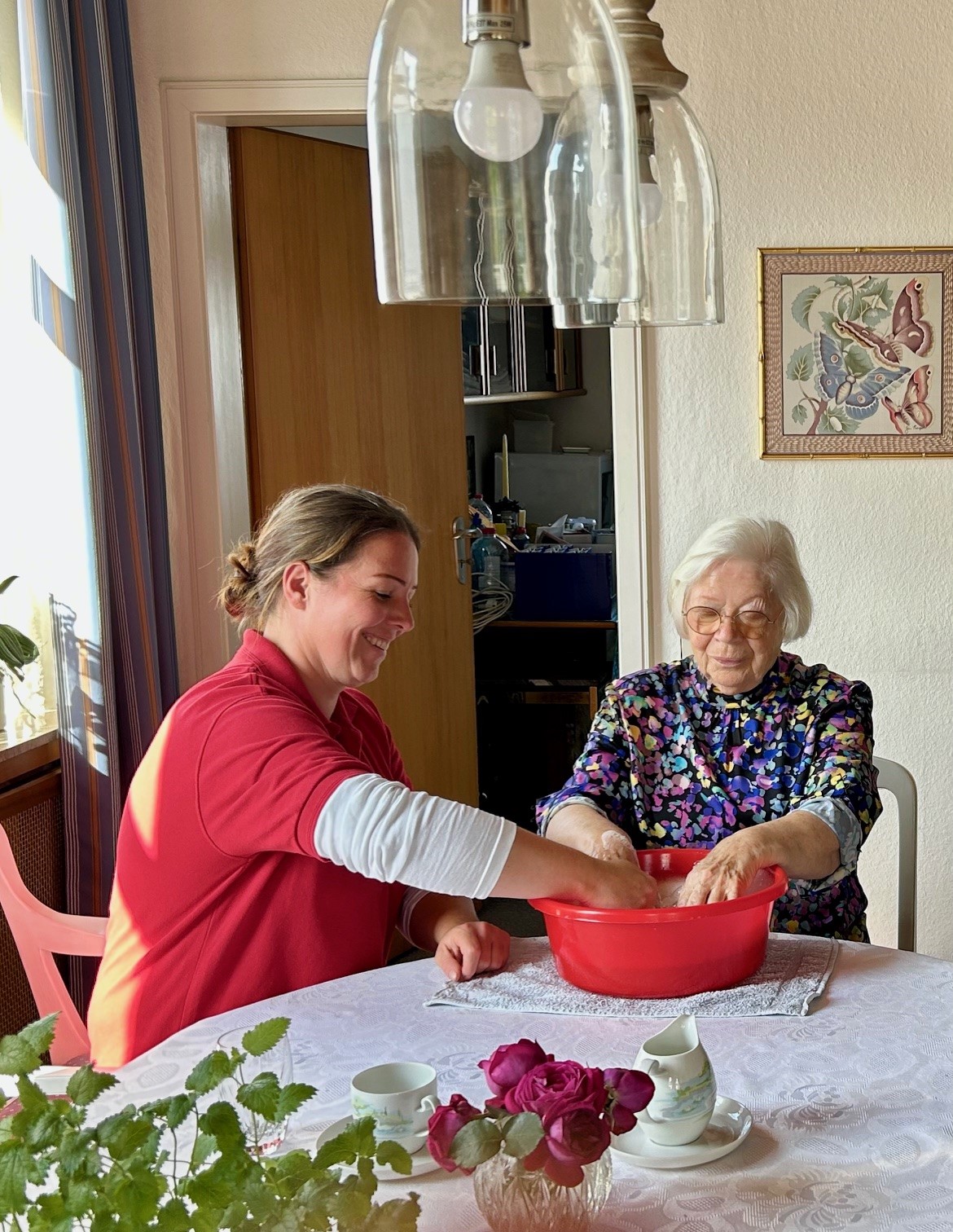 Pflegekraft und ältere Frau waschen gemeinsam die Hände in einer roten Schüssel auf einem gedeckten Tisch.