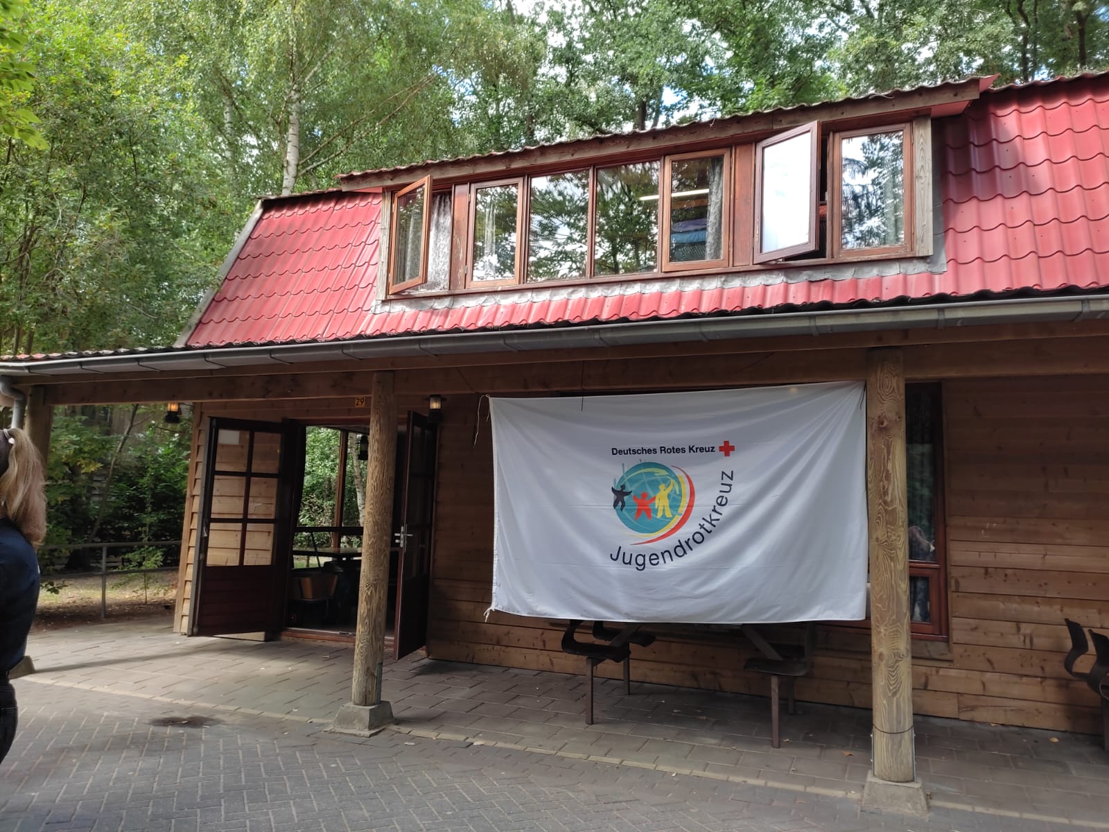 Holzgebäude mit rotem Ziegeldach und offenen Türen, an dessen Front eine weiße Jugendrotkreuz-Flagge mit farbigem Logo hängt.