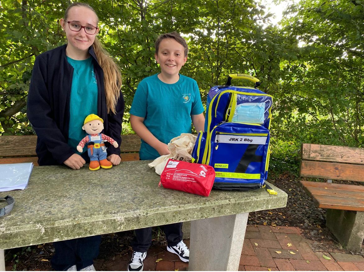 Eine Frau und ein Junge stehen hinter einem Steintisch mit blauem Notfallrucksack, roter Erste-Hilfe-Tasche und einer Bob-der-Baumeister-Figur, im Freien unter grünen Bäumen.