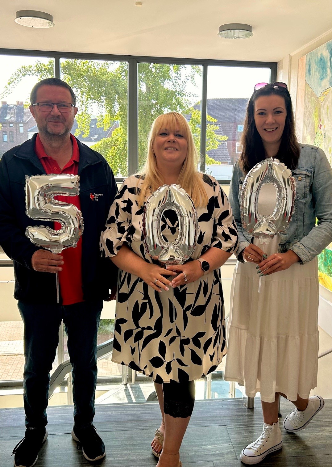 Drei Personen halten silberne Luftballons in der Form der Zahl 500 vor einem Fenster mit Grün im Hintergrund.