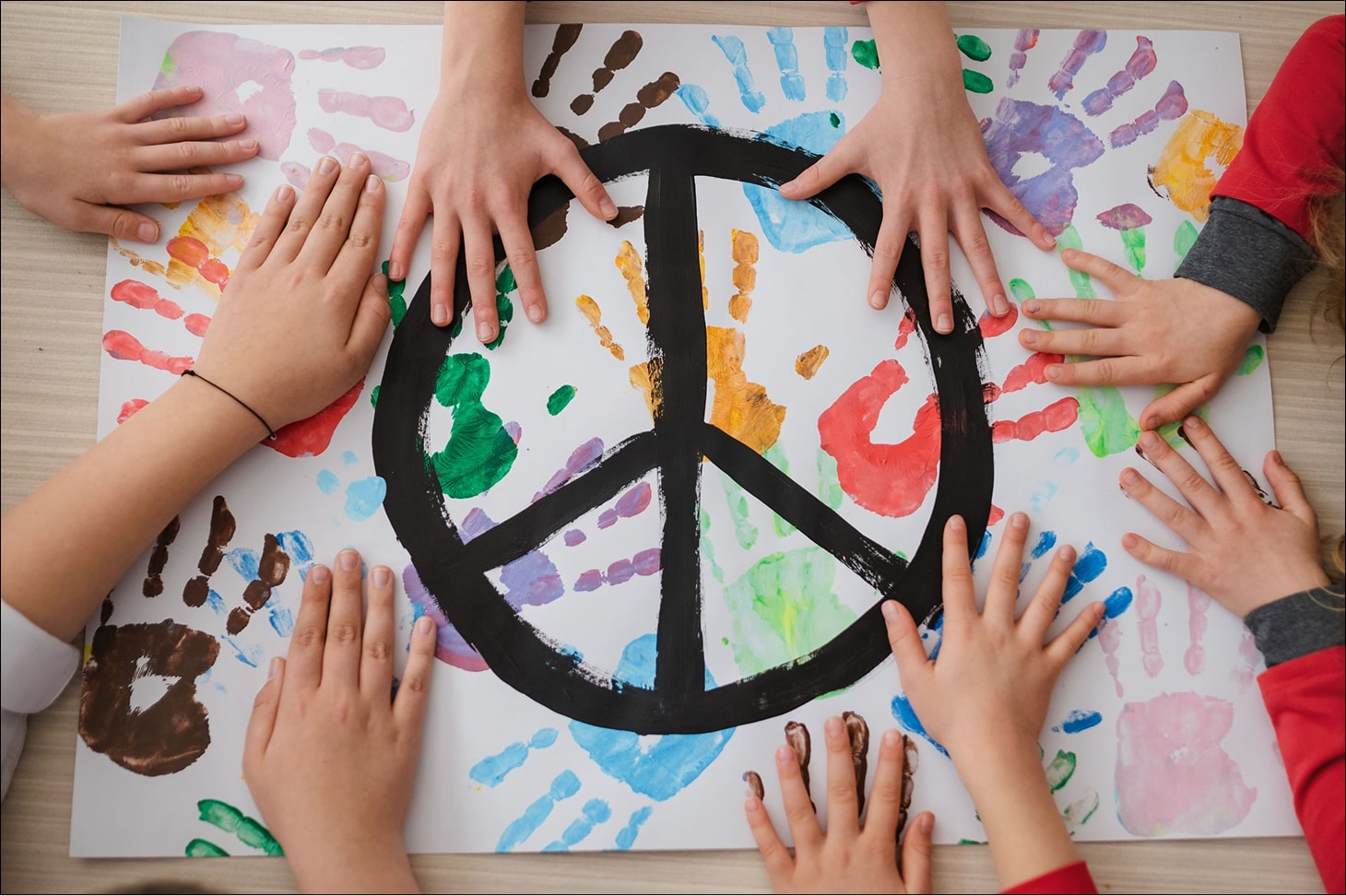 Kinderhände liegen um ein Blatt Papier mit bunten Handabdrücken und einem schwarzen Friedenssymbol in der Mitte.