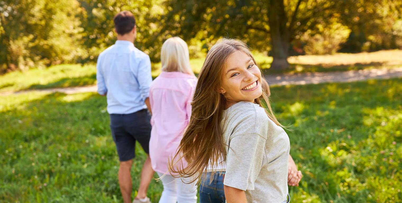 Lächelndes Mädchen schaut über die Schulter zurück, während ihre Eltern voraus auf einem sonnigen Parkweg gehen.