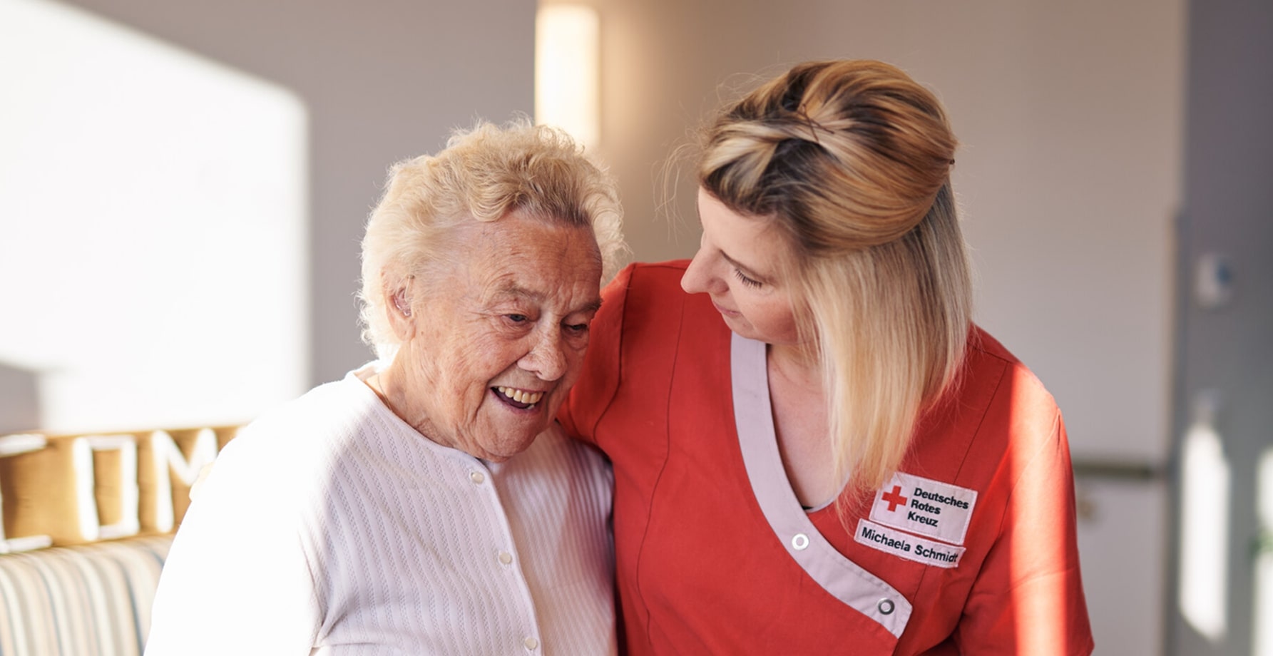 Ältere Frau lächelt und wird von einer weiblichen Pflegekraft in roter Uniform drinnen unterstützt.