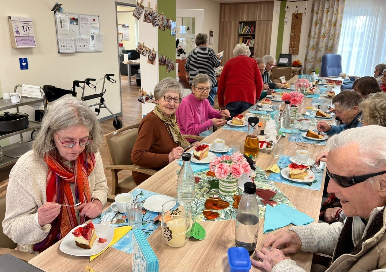 Gruppe älterer Menschen sitzt an einem langen Tisch und genießt Kaffee und Kuchen in einem hellen Raum, der mit Blumen und Fotos dekoriert ist.