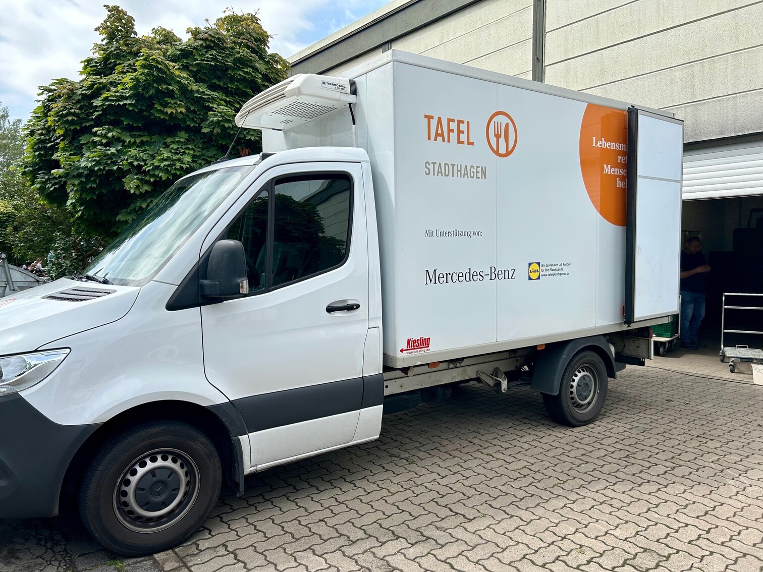 Weißer Kühltransporter mit Tafel-Stadthagen-Beschriftung, geparkt an einem Gebäude, mit einer Person im Inneren.