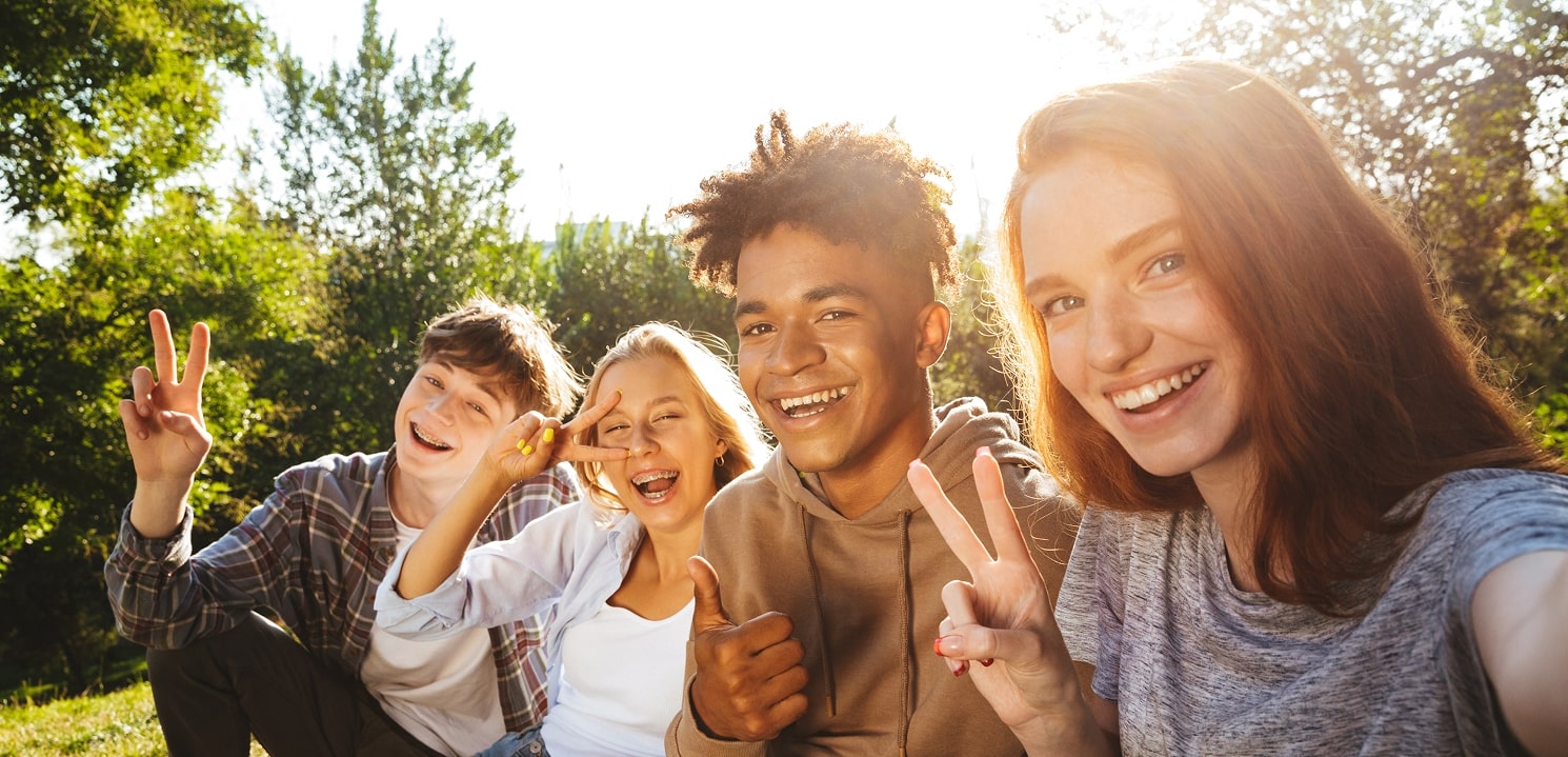 Vier Jugendliche im Freien lächeln und zeigen Peace-Zeichen und Daumen hoch bei Sonnenlicht.